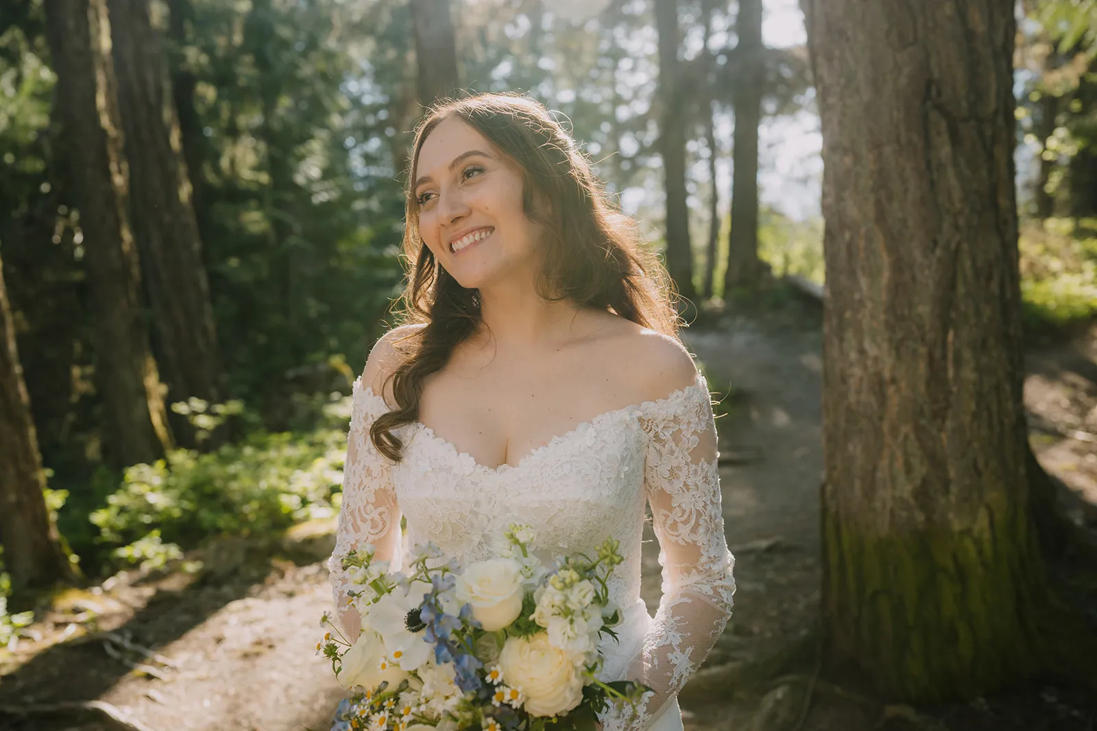 Bruna smiling up into golden dappled sunlight on a forest trail, holding a bouquet of white roses, blue delphiniums, and wild flowers