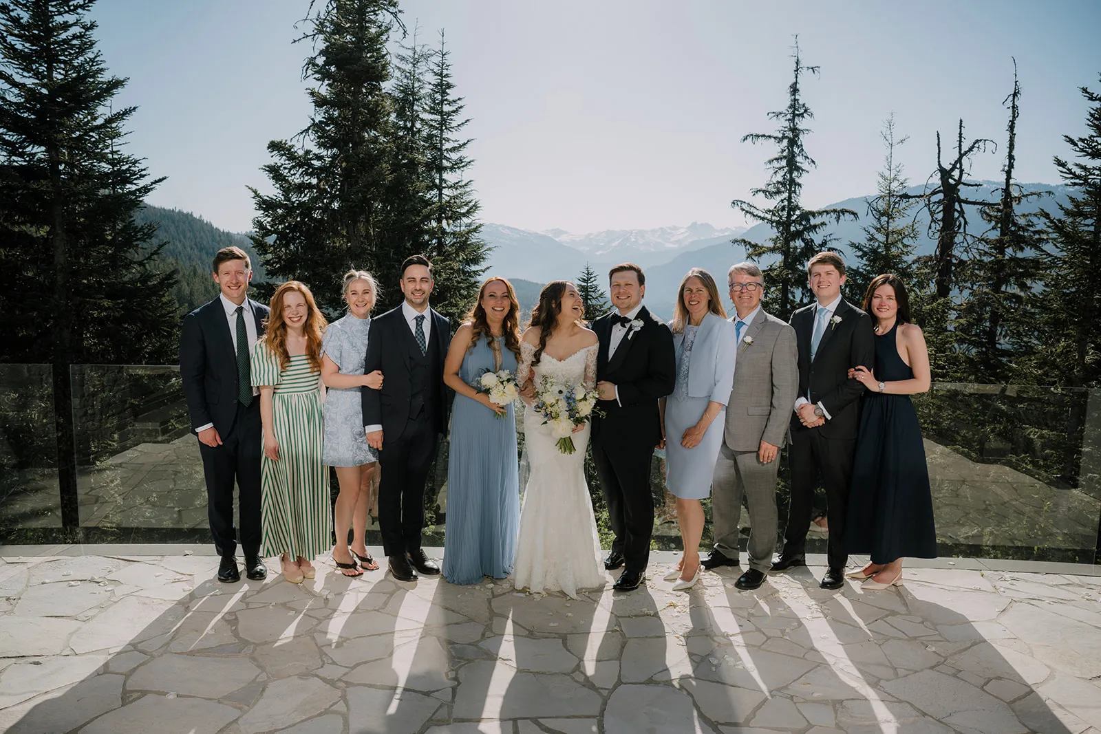 Group photo of the whole party on a stone terrace, with snow-capped mountains and pine forest behind them