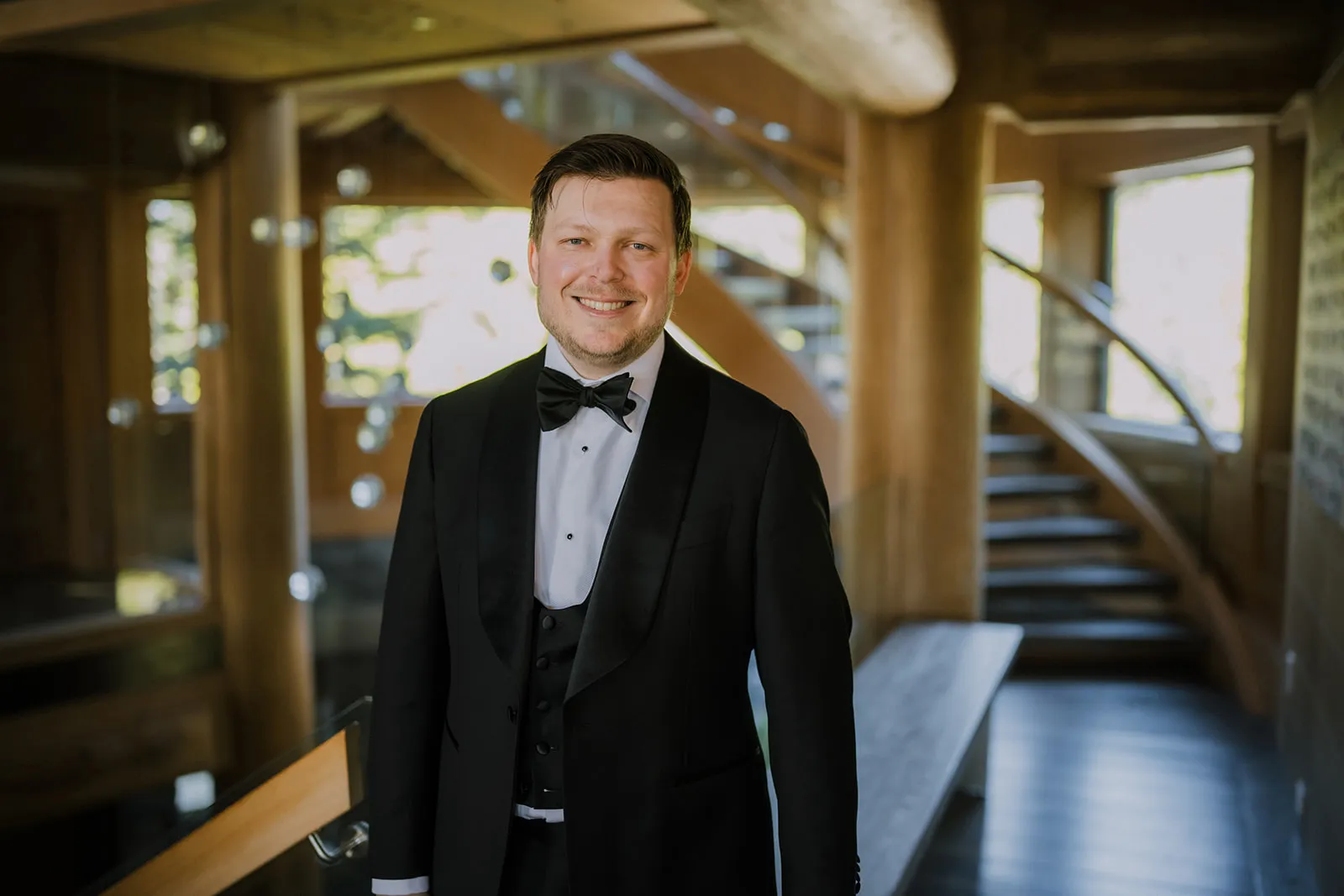 James smiling in his black tuxedo and bow tie inside the timber-framed lodge, the sweeping staircase visible behind him
