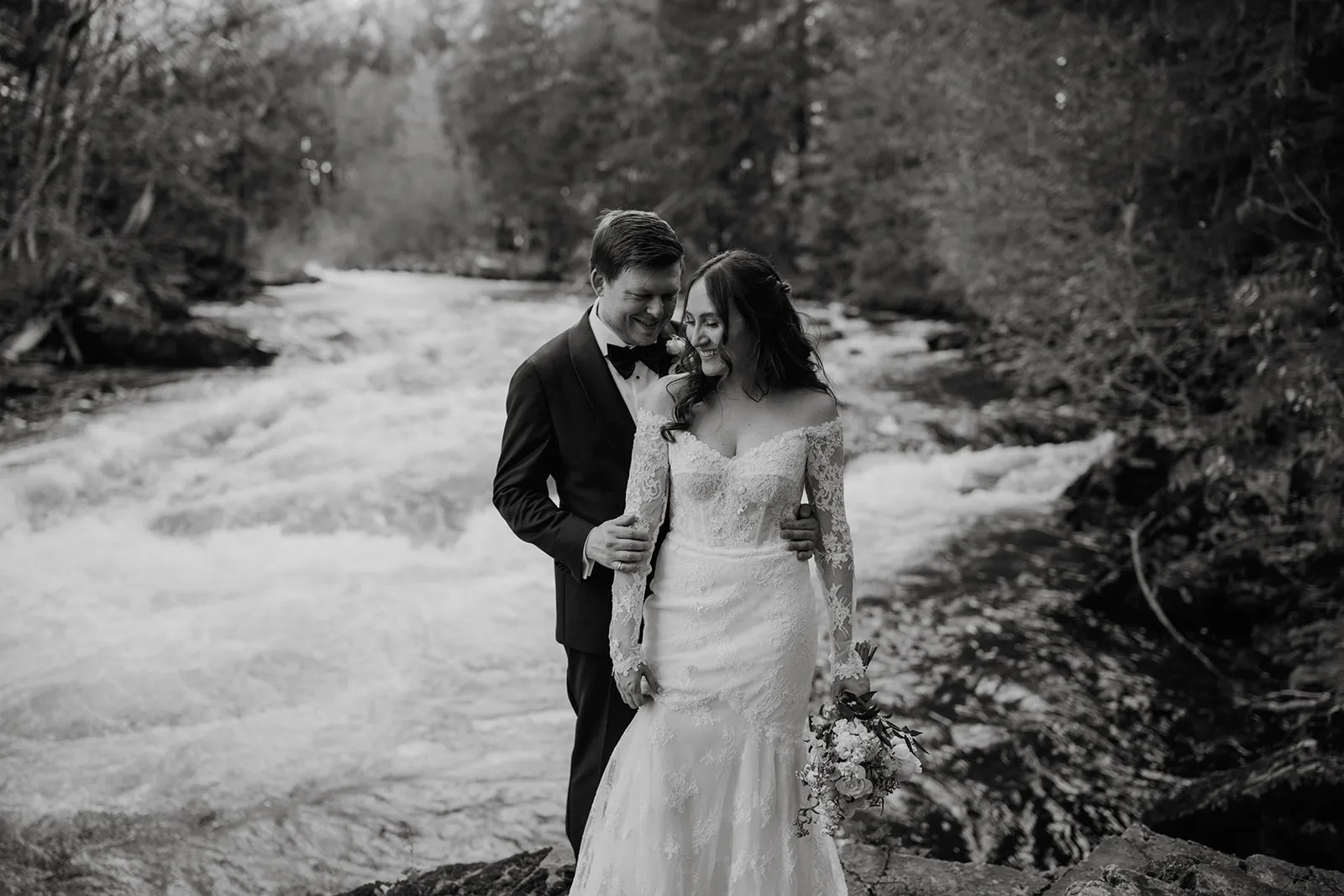 Black and white — James nuzzling Bruna's cheek as they both grin, standing beside a fast-flowing glacial river in the Whistler forest