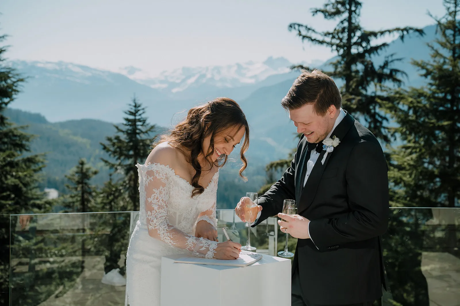 Both laughing as Bruna signs the register on an outdoor terrace, champagne in hand, with the snow-capped Whistler mountains behind them