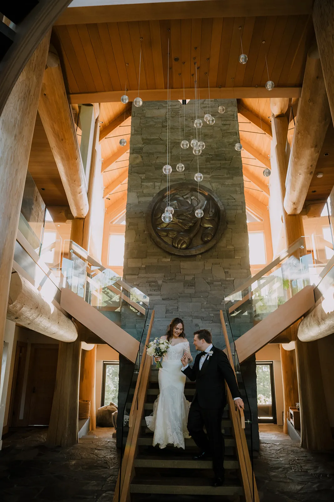 James leads Bruna down a sweeping staircase inside the timber-framed lodge, laughing together beneath a cascade of hanging glass globe lights