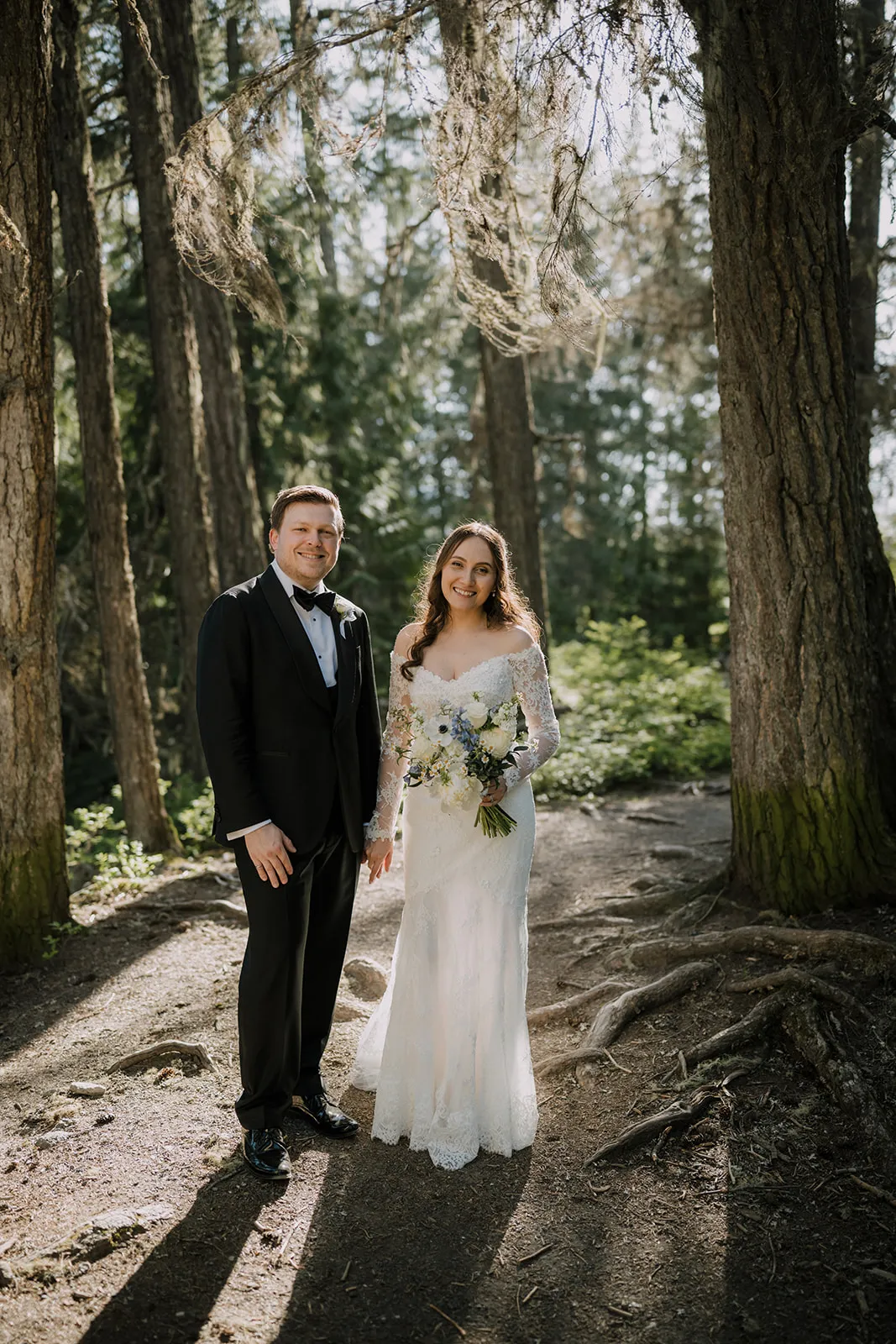 James and Bruna standing together on a forest path between tall pine trees, Bruna in a lace off-the-shoulder gown holding a white-and-blue bouquet, James in a black tuxedo